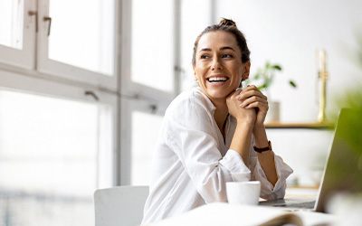 Smiling woman in white shirt at desk with laptop in front of windows